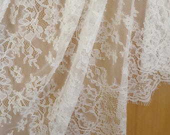 May include: Close-up of white lace fabric with a floral pattern. The delicate lace is draped, revealing intricate details and a scalloped edge. The fabric is likely used for apparel or home decor.