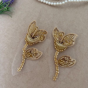 May include: Two gold flower-shaped patches featuring intricate beadwork and sequins on a sheer mesh backing. Each patch depicts a stylized blossom with a stem and a single leaf, crafted with metallic gold beads and shimmering sequins.