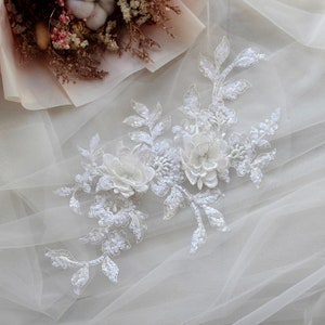 Puede incluir: Aplique floral blanco con lentejuelas y cuentas. El aplique presenta dos flores grandes y hojas delicadas. Este adorno es perfecto para agregar un toque de elegancia a los vestidos de novia, los accesorios de novia u otras prendas para ocasiones especiales.