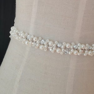 May include: A white beaded sash featuring pearl and crystal embellishments. The sash is ideal for adding a touch of elegance to a wedding dress or other formal attire.