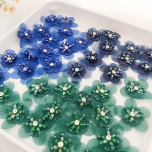 May include: Assortment of blue and green fabric flower embellishments with rhinestone centres. The flowers are arranged on a white surface.