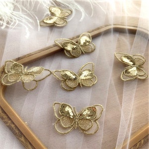 May include: Six gold butterfly-shaped fabric appliques with sequins. The appliques are on a white mesh background.