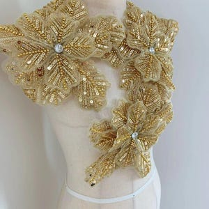 Könnte beinhalten: Goldene Perlen- und Pailletten-Blumenapplikation mit Strassstein in der Mitte. Diese Verzierung ist perfekt, um jedem Kleidungsstück einen Hauch von Glanz und Eleganz zu verleihen.