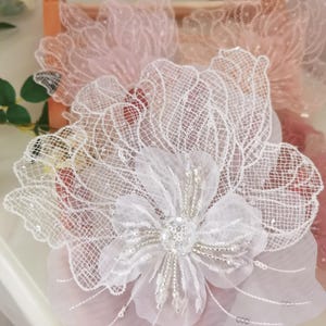 May include: White lace flower applique with beaded center. The flower is delicate and intricate, with a soft, flowing design.