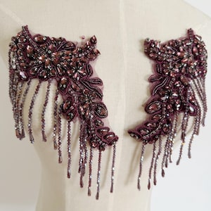 May include: A pair of symmetrical dark purple beaded appliques featuring intricate floral-inspired embroidery, faceted crystals, and dangling beaded fringe strands. These decorative embellishments are crafted on a sheer mesh base for attachment.