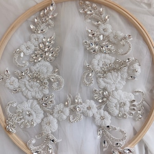 May include: Two white lace appliques with silver rhinestone embellishments. The appliques are displayed on a wooden embroidery hoop.