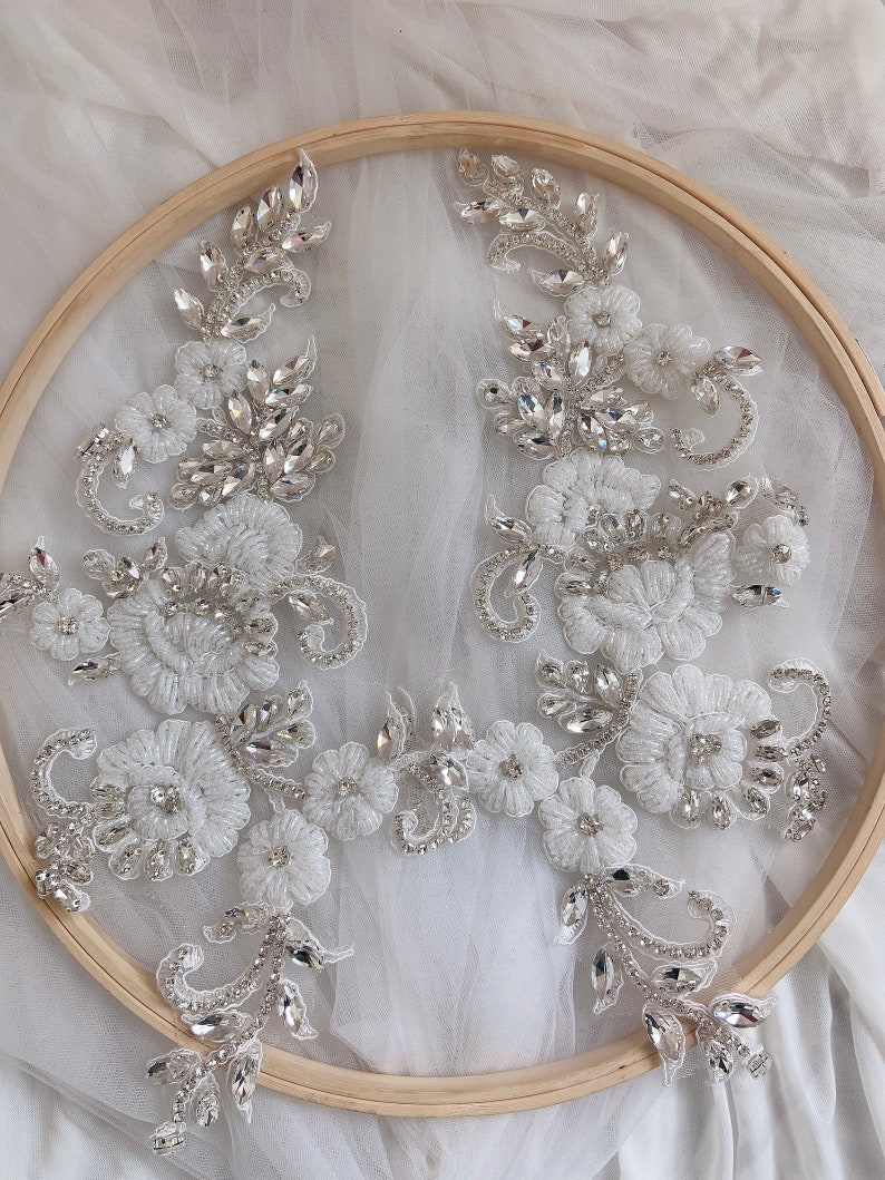 May include: Two white lace appliques with silver rhinestone embellishments. The appliques are displayed on a wooden embroidery hoop.