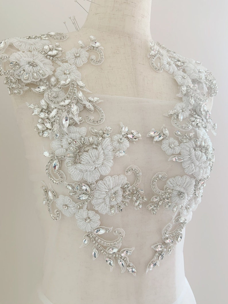 May include: White lace applique with crystal embellishments. The applique features floral designs and intricate details. This applique is perfect for adding a touch of elegance to any garment.