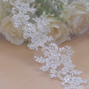 Puede incluir: Borde de encaje blanco con diseños florales y lentejuelas. El borde está sobre una tela blanca transparente.