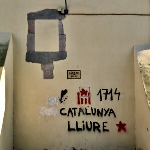 以下が含まれることがあります： 「CATALUNYA LLIURE」と数字の「1714」を含む、黒と赤の落書きが施された壁。赤い星と旗のようなシンボルも描かれています。街灯も見えます。