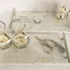 May include: Two white marble trays with silver handles. The top tray has a silver coffee pot, a glass, and a small box with the text "Damat Kahvesi". The bottom tray has two glass boxes with white roses, a pair of silver scissors, and a silver tassel. The trays are engraved with the names "Ceren & Semih" and "Semih'in Damat Kahvesi".