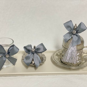 Può includere: Tre pezzi da servizio in argento con fiocchi di nastro grigi e un vassoio in argento. I pezzi da servizio sono una piccola tazza di vetro, una piccola tazza d'argento e una tazza d'argento più grande con manico e piattino.