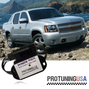 May include: A black power pack with the text "POWER PACK QUICK CONNECT TURBO TUNER PROTUNINGUSA WWW.PROTUNINGUSA.COM" on it. The power pack is sitting on a rocky surface in front of a silver pickup truck.