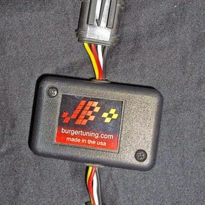 May include: A black electronic device with a red and yellow logo that says "burgertuning.com made in the usa". The device has four wires connected to it, two on each side.