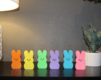 Pode incluir: Uma fila de sete figurinhas de coelho coloridas, cada uma com uma cor diferente: laranja, amarelo, verde, roxo, azul e rosa. As figurinhas estão dispostas em linha sobre uma superfície preta.