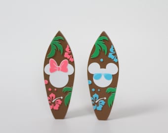 May include: Two brown surfboard-shaped ornaments with a tropical theme. One features a white silhouette of Mickey Mouse with blue sunglasses and the other features a white silhouette of Minnie Mouse with a pink bow.