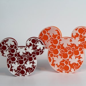 May include: Three decorative Mickey Mouse head-shaped cutouts. One is a deep burgundy color, and two are orange. Each cutout features a white maple leaf and Mickey Mouse head pattern. The cutouts are likely for fall or autumn-themed decor.