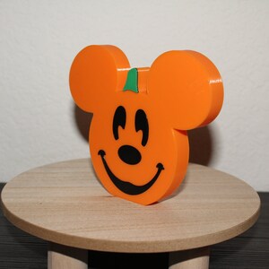 May include: Orange pumpkin-shaped Mickey Mouse figurine with a green stem and a black smiling face.