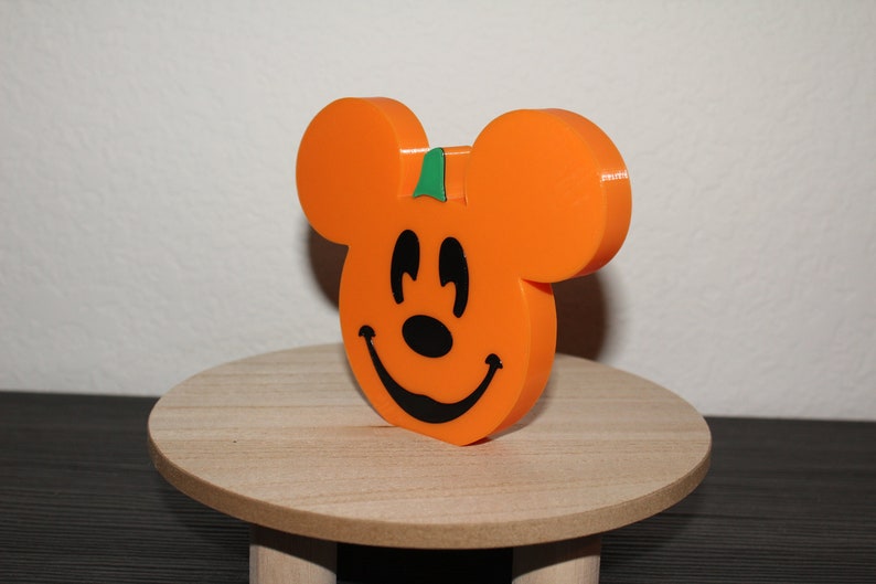 May include: Orange pumpkin-shaped Mickey Mouse figurine with a green stem and a black smiling face.