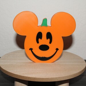 May include: Orange pumpkin shaped like Mickey Mouse with a green stem and a black smiling face.