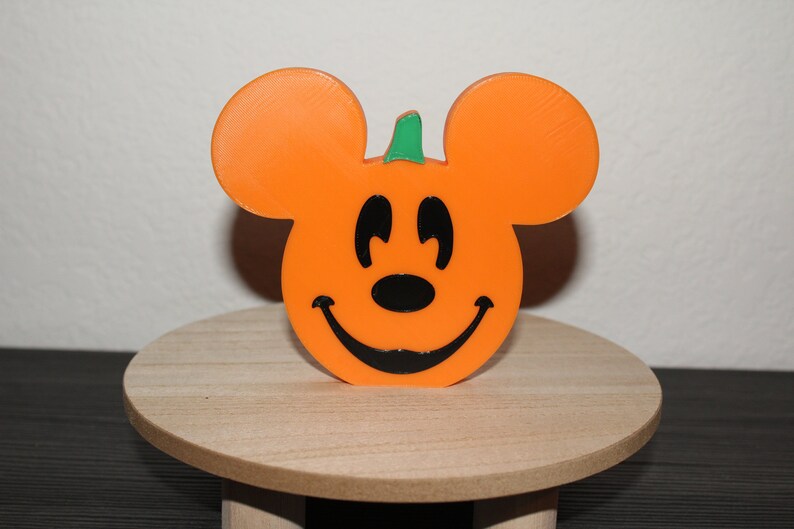 May include: Orange pumpkin shaped like Mickey Mouse with a green stem and a black smiling face.