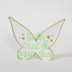 May include: White butterfly-shaped wooden sign with gold trim and gold glitter stars. The sign says "faith trust & pixie dust" in green script.