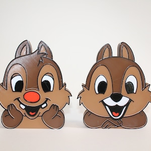 May include: Two brown chipmunk figurines with white eyes and black pupils. The chipmunks are smiling with their mouths open and have red noses. One chipmunk is holding its hands up to its cheeks and the other is resting its chin on its hands.