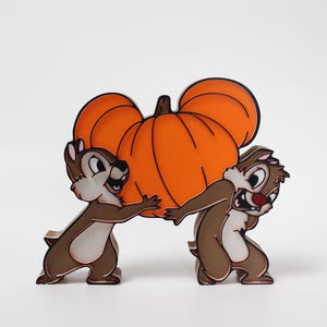 May include: A decorative figurine featuring Chip and Dale, Disney characters, struggling to carry a large orange pumpkin. The pumpkin has a Mickey Mouse-inspired design. The figurine is brown, orange, and white.
