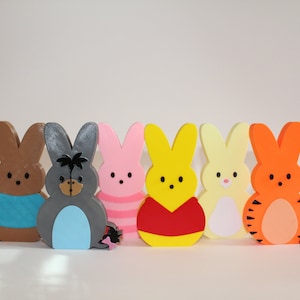May include: A set of six colorful bunny-shaped figurines, each with a different design. The bunnies are standing in a row on a white surface. The bunnies are made of wood and painted in various colors, including brown, gray, pink, yellow, white, and orange.