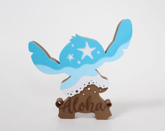May include: A blue and white wooden cutout of Stitch from Lilo & Stitch, with a wave design and a star. The cutout is standing on a brown wooden base with the word "Aloha" carved into it.