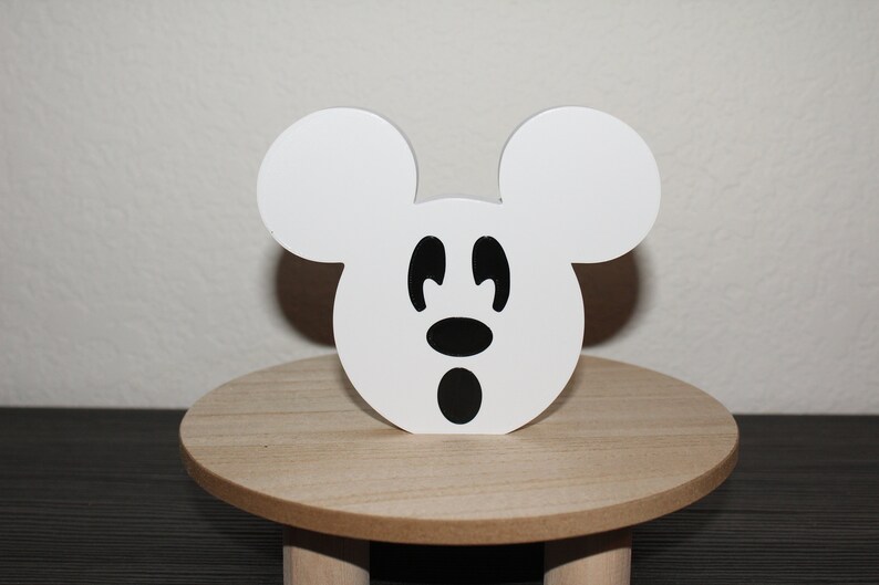 May include: White wooden Mickey Mouse head with black eyes, nose, and mouth. The head is sitting on a wooden stand.