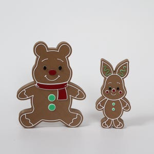 May include: Two gingerbread cookie ornaments, one shaped like a bear and the other shaped like a rabbit. Both are decorated with white icing and green and red accents.