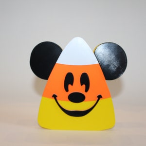 May include: A yellow, orange, and white candy corn shaped ornament with a black Mickey Mouse face. The ornament has black ears on either side.