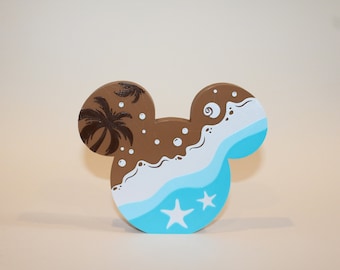 May include: A wooden Mickey Mouse head painted with a beach scene. The left ear is painted brown with a palm tree and white bubbles. The right ear is painted blue with white waves, a seashell, and white stars.