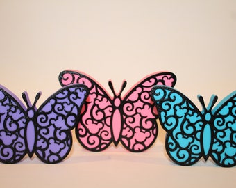 May include: Three decorative butterflies made of wood with intricate black filigree designs. The butterflies are painted in shades of purple, pink, and blue.