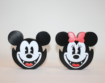 Puede incluir: Un par de figuras impresas en 3D en blanco y negro de Mickey Mouse y Minnie Mouse con colmillos, como si fueran vampiros. Mickey Mouse está a la izquierda y Minnie Mouse a la derecha. Minnie Mouse tiene un lazo rojo en la cabeza.