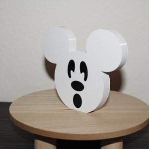 May include: White wooden Mickey Mouse head cutout with black eyes and a black nose and mouth. The cutout is sitting on a small wooden pedestal.