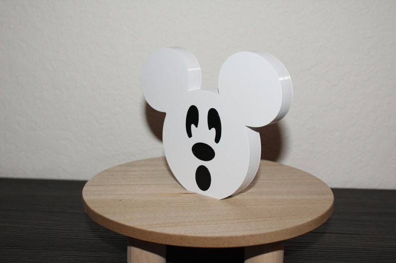May include: White wooden Mickey Mouse head cutout with black eyes and a black nose and mouth. The cutout is sitting on a small wooden pedestal.
