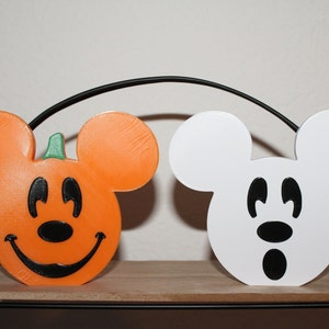 May include: Two Halloween-themed Mickey Mouse figures. One is a pumpkin-shaped Mickey Mouse with a smiling face and the other is a white Mickey Mouse with a surprised expression.