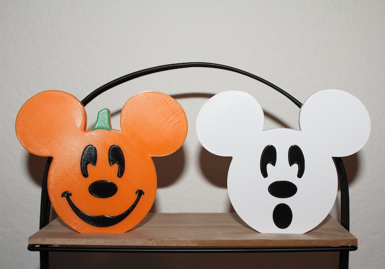 May include: Two Halloween-themed Mickey Mouse figures. One is a pumpkin-shaped Mickey Mouse with a smiling face and the other is a white Mickey Mouse with a surprised expression.