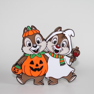 May include: A decorative enamel pin featuring two brown chipmunks in Halloween costumes. One chipmunk wears an orange pumpkin suit with a matching hat and carries a small pumpkin bucket. The other wears a white ghost sheet and a striped scarf.