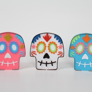 May include: Three colorful sugar skull decorations with intricate floral designs and bright colors. The skulls are pink, white, and blue.