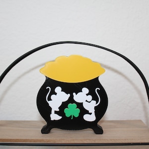 May include: A black pot with a yellow lid and white silhouettes of Mickey Mouse and Minnie Mouse holding a green shamrock. The pot is sitting on a wooden shelf.