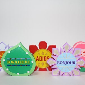 May include: Five colorful wooden flower-shaped decorations with different greetings in different languages. The decorations are green, red, yellow, purple, and pink. The greetings are "Kwaheri", "Adios", "Bonjour", "Will you", and "さようなら".