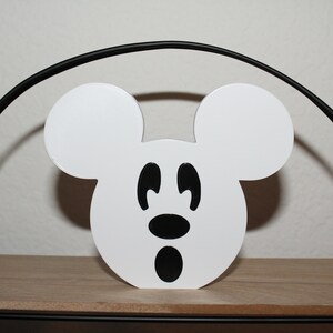 May include: White wooden cutout of Mickey Mouse's head with black eyes, nose, and mouth. The cutout is sitting on a wooden shelf.
