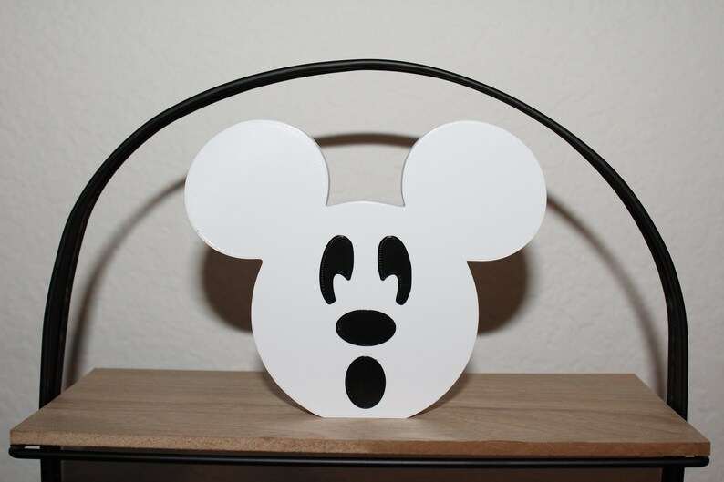 May include: White wooden cutout of Mickey Mouse's head with black eyes, nose, and mouth. The cutout is sitting on a wooden shelf.