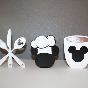 May include: Three white wooden figurines with black details. The first figurine is a set of crossed utensils, a fork, knife, and spoon, with Mickey Mouse ears on the handles. The second figurine is Mickey Mouse wearing a chef's hat. The third figurine is a coffee mug with Mickey Mouse ears on the side.