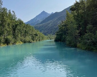 Pode incluir: Uma paisagem cénica com um rio turquesa serpenteando por um vale. Árvores verdes exuberantes margeiam as margens, com uma cordilheira ao fundo sob um céu azul claro. A cor da água contrasta com a vegetação circundante.