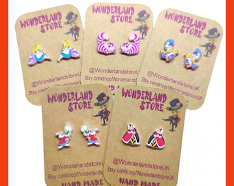 Op de afbeelding: Zes paar handgemaakte Alice in Wonderland-oorbellen op bruin karton met de tekst "Wonderland Store" en social media-handvatten. De oorbellen bevatten personages zoals de Koningin van harten, de Hoedenmaker, het Witte Konijn en de Cheshire Cat.
