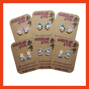 May include: Four cards with the text "Wonderland Store" on them. Each card features a pair of small, colourful, cartoon character earrings. The characters are Donald Duck and Daisy Duck. The cards are on a brown background. The text "Hand Made" is on the bottom of each card.